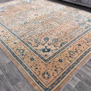 Op de afbeelding: Een vintage tapijt met een beige achtergrond en een blauw en wit bloemenpatroon. Het tapijt heeft een rand met een herhalend patroon van blauw en wit.