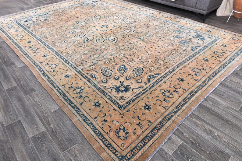 Op de afbeelding: Een vintage tapijt met een beige achtergrond en een blauw en wit bloemenpatroon. Het tapijt heeft een rand met een herhalend patroon van blauw en wit.
