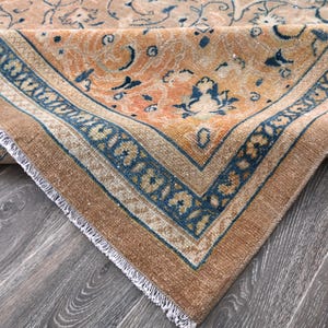 Op de afbeelding: Close-up van een vintage tapijt met een vervaagd oranje en blauw bloemenpatroon. Het tapijt heeft een gefranst randje en is dubbelgevouwen.