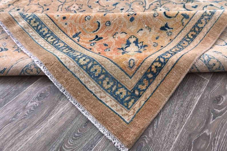 Op de afbeelding: Close-up van een vintage tapijt met een vervaagd oranje en blauw bloemenpatroon. Het tapijt heeft een gefranst randje en is dubbelgevouwen.
