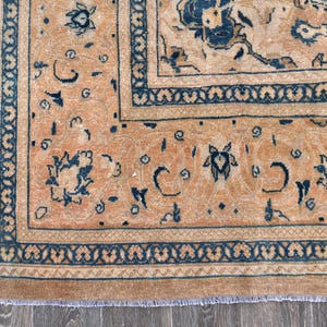 Op de afbeelding: Een vintage tapijt met een perzikkleurige achtergrond en een donkerblauw bloemenpatroon. Het tapijt heeft een franje rand.