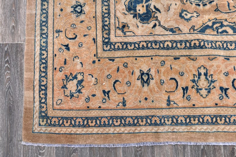 Op de afbeelding: Een vintage tapijt met een perzikkleurige achtergrond en een donkerblauw bloemenpatroon. Het tapijt heeft een franje rand.