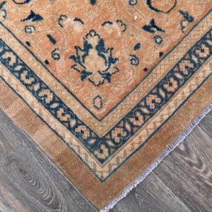 Op de afbeelding: Een vintage tapijt met een verbleekte oranje achtergrond en een donkerblauw geometrisch patroon. Het tapijt heeft een gefranst randje.