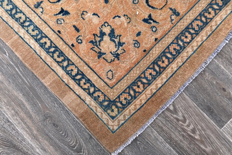 Op de afbeelding: Een vintage tapijt met een verbleekte oranje achtergrond en een donkerblauw geometrisch patroon. Het tapijt heeft een gefranst randje.