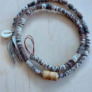 Puede incluir: Un cordón trenzado marrón, blanco y gris con un pequeño amuleto de concha blanca y una cuenta de madera.