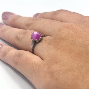 Op de afbeelding: Een ring in vintage-stijl met een grote, ovale roze edelsteen. De ring heeft een gedetailleerde, sierlijke zilveren band met kleine accentstenen. De edelsteen is het middelpunt, gezet in een decoratieve setting.