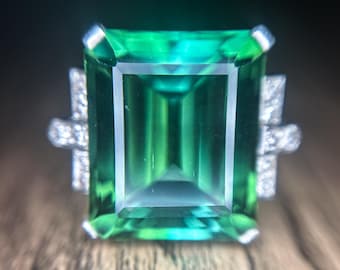 May include: A large, emerald-cut green gemstone ring with a rectangular center stone. The ring features small, sparkling accent stones on either side of the main stone, set in a silver or white metal band. The gemstone has a vibrant, transparent appearance.