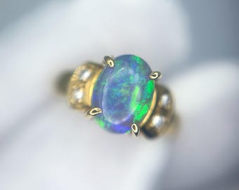 Op de afbeelding: Een gouden ring met een ovale opaal edelsteen. De opaal vertoont een levendig kleurenspel, met blauwe en groene flitsen. De ring heeft een klassiek ontwerp met kleine accentstenen op de band.