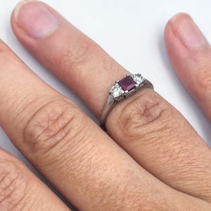 May include: A silver ring featuring a rectangular, deep red gemstone flanked by two smaller, round, clear gemstones. The ring is set on a finger against a white background, showcasing the jewelry's design and color contrast.
