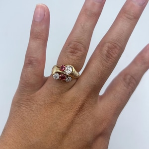 May include: A gold bypass ring featuring two round-cut red gemstones and two round-cut clear white diamonds. The band has a textured, ribbed design that wraps around the finger, creating a layered look with the four stones set in a diagonal pattern.