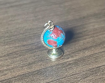 May include: A miniature globe pendant with a silver-colored frame and base. The globe features a blue ocean and red landmasses. The pendant is designed to be worn as a necklace charm.