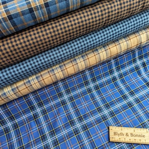 May include: Four different plaid fabrics stacked on top of each other. The top fabric is a blue and brown plaid, the second fabric is a brown and tan gingham, the third fabric is a blue and tan gingham, and the bottom fabric is a blue, brown, and tan plaid. A wooden ruler with the text "Blyth & Bonnie" is on top of the fabric.