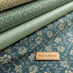 May include: A stack of fabric rolls in shades of teal and cream. The top roll features a small, repeating pattern, while the bottom roll displays a floral design. A wooden ruler with the text "Blyth & Bonnie" rests on the bottom fabric.