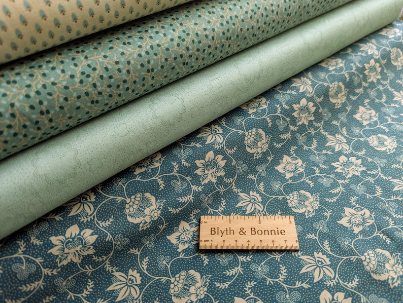 Puede incluir: Una pila de rollos de tela en tonos turquesa y crema. El rollo superior presenta un peque&ntilde;o patr&oacute;n repetitivo, mientras que el rollo inferior muestra un dise&ntilde;o floral. Una regla de madera con el texto "Blyth & Bonnie" descansa sobre la tela inferior.