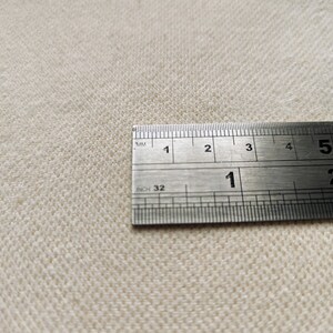 May include: Close-up of a beige fabric with a stainless steel ruler. The ruler is marked in inches and millimeters, showing measurements up to 15 cm. The fabric has a textured weave.