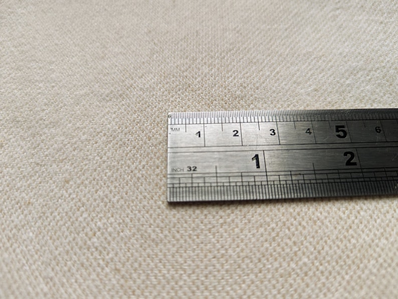May include: Close-up of a beige fabric with a stainless steel ruler. The ruler is marked in inches and millimeters, showing measurements up to 15 cm. The fabric has a textured weave.
