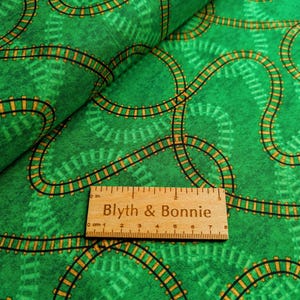 Puede incluir: Tela verde con un patrón repetido de vías de tren negras y amarillas. Una regla de madera con el texto "Blyth & Bonnie" está colocada sobre la tela. La tela tiene una apariencia texturizada.