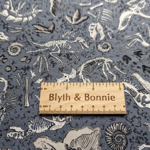 May include: Gray fabric with a white dinosaur skeleton print. The fabric features a variety of dinosaur skeletons, including a T-Rex, a Triceratops, and a Stegosaurus. The fabric also features fossilized footprints and other prehistoric artifacts. The fabric is 7 inches wide.