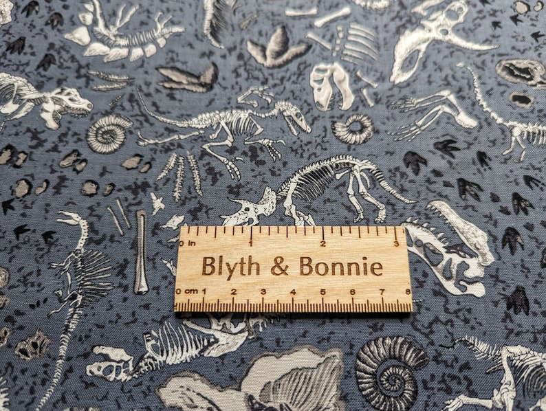 May include: Gray fabric with a white dinosaur skeleton print. The fabric features a variety of dinosaur skeletons, including a T-Rex, a Triceratops, and a Stegosaurus. The fabric also features fossilized footprints and other prehistoric artifacts. The fabric is 7 inches wide.