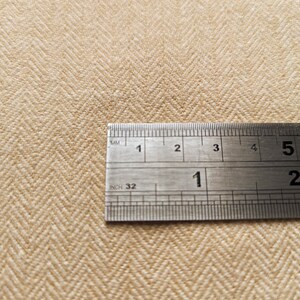 May include: Close-up of beige herringbone fabric with a metal ruler showing inch and millimeter measurements. The fabric has a textured appearance and a diagonal pattern. The ruler is positioned across the fabric, providing a size reference.