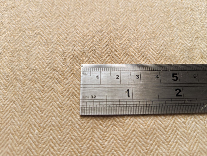 May include: Close-up of beige herringbone fabric with a metal ruler showing inch and millimeter measurements. The fabric has a textured appearance and a diagonal pattern. The ruler is positioned across the fabric, providing a size reference.