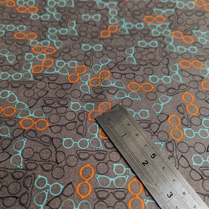 May include: Gray fabric with a repeating pattern of eyeglasses in black, orange, and teal. A metal ruler is placed diagonally across the fabric, showing measurements in inches.
