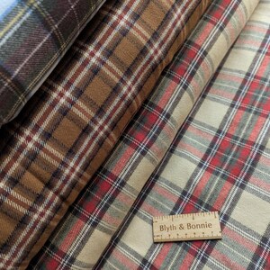 May include: Close-up of several rolls of plaid fabric in various colours. The fabrics feature a classic plaid pattern in shades of brown, red, cream, and blue. A wooden ruler with the text "Blyth & Bonnie" is visible.