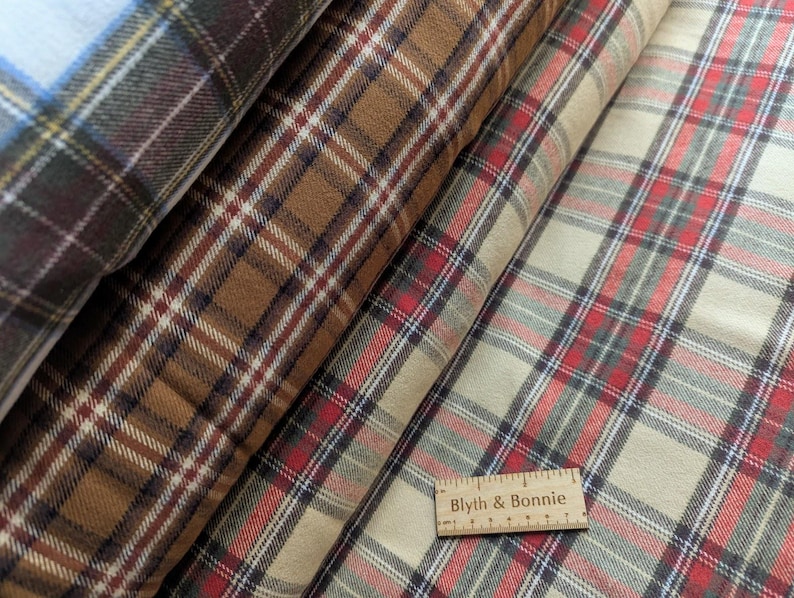 May include: Close-up of several rolls of plaid fabric in various colours. The fabrics feature a classic plaid pattern in shades of brown, red, cream, and blue. A wooden ruler with the text "Blyth & Bonnie" is visible.