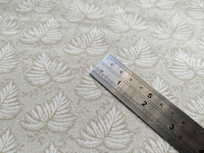 Pode incluir: Um close-up de um tecido bege com um padr&atilde;o de folhas brancas. Uma r&eacute;gua de metal &eacute; colocada em cima do tecido, mostrando medidas em polegadas.