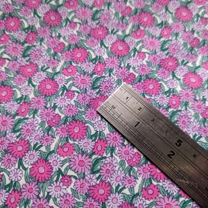 Pode incluir: Um tecido branco com um padr&atilde;o repetitivo de flores rosa e roxas. Uma r&eacute;gua de metal &eacute; colocada em cima do tecido.