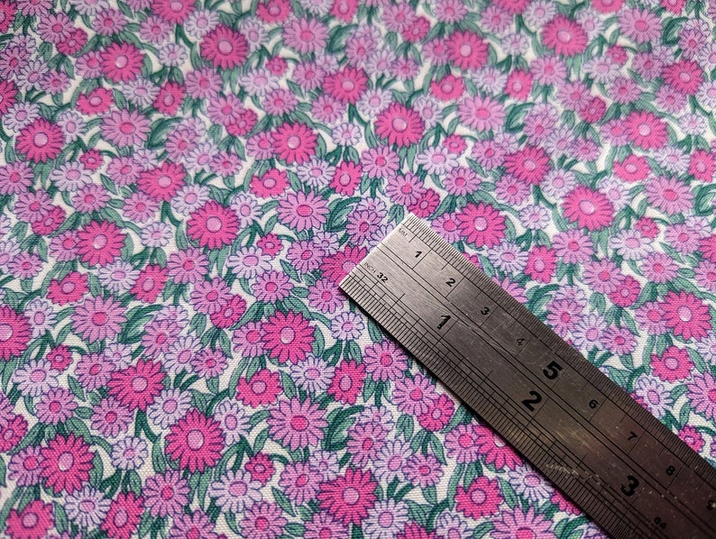 Pode incluir: Um tecido branco com um padr&atilde;o repetitivo de flores rosa e roxas. Uma r&eacute;gua de metal &eacute; colocada em cima do tecido.