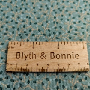 Puede incluir: Una regla de madera con medidas en pulgadas y cent&iacute;metros, etiquetada como "Blyth & Bonnie", descansa sobre una tela verde azulado con un patr&oacute;n repetido de peque&ntilde;os puntos verde oscuro y azul claro y ramas beige. La regla mide aproximadamente 7,6 cm de largo.