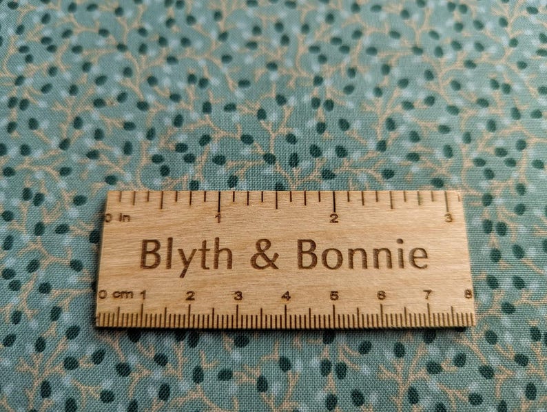 Puede incluir: Una regla de madera con medidas en pulgadas y cent&iacute;metros, etiquetada como "Blyth & Bonnie", descansa sobre una tela verde azulado con un patr&oacute;n repetido de peque&ntilde;os puntos verde oscuro y azul claro y ramas beige. La regla mide aproximadamente 7,6 cm de largo.