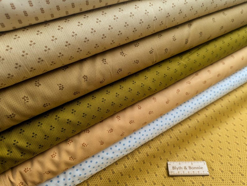 Puede incluir: Una pila de rollos de tela en varios colores, incluyendo beige, verde oliva y amarillo. Cada tela tiene un peque&ntilde;o patr&oacute;n floral repetido. Las telas est&aacute;n dispuestas en diagonal, con una regla de madera "Blyth & Bonnie" en la parte inferior.