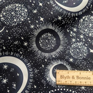 May include: Dark navy fabric with a celestial pattern featuring white crescent moons, stars, and sunbursts. A wooden ruler with the text "Blyth & Bonnie" is in the lower right corner. The fabric has a cosmic, space-themed design.