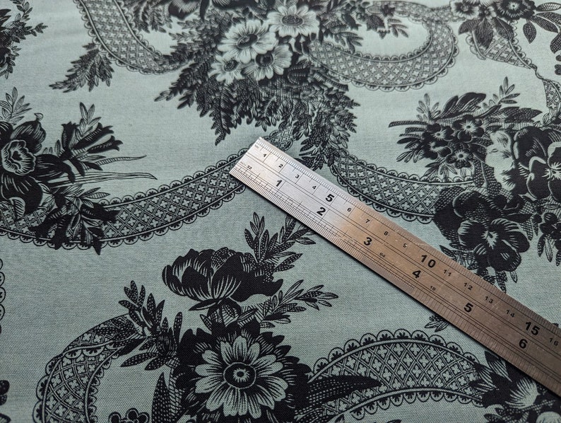 Pode incluir: Um close-up de um tecido turquesa com um padr&atilde;o floral preto. O tecido tem um padr&atilde;o repetitivo de flores e folhas. Uma r&eacute;gua &eacute; colocada em cima do tecido, mostrando a escala.