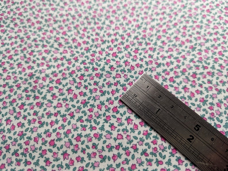 Pode incluir: Tecido branco com um padr&atilde;o repetitivo de pequenas flores cor-de-rosa com folhas verdes. Uma r&eacute;gua de metal &eacute; colocada sobre o tecido, mostrando a escala em cent&iacute;metros.