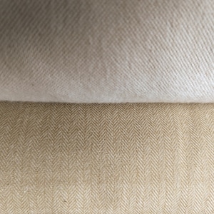 May include: Close-up of two fabric swatches. The top swatch is a solid off-white colour, while the bottom swatch has a tan herringbone pattern. The fabrics are layered, showcasing the textures and colours.