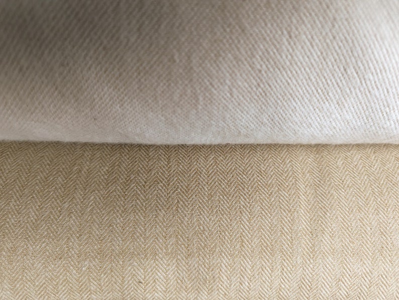 May include: Close-up of two fabric swatches. The top swatch is a solid off-white colour, while the bottom swatch has a tan herringbone pattern. The fabrics are layered, showcasing the textures and colours.