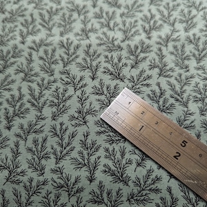Pode incluir: Um close-up de um tecido verde com um padr&atilde;o repetitivo de pequenos ramos pretos estilizados. Uma r&eacute;gua de metal &eacute; colocada sobre o tecido, com os n&uacute;meros 1, 2, 3 e 4 vis&iacute;veis.