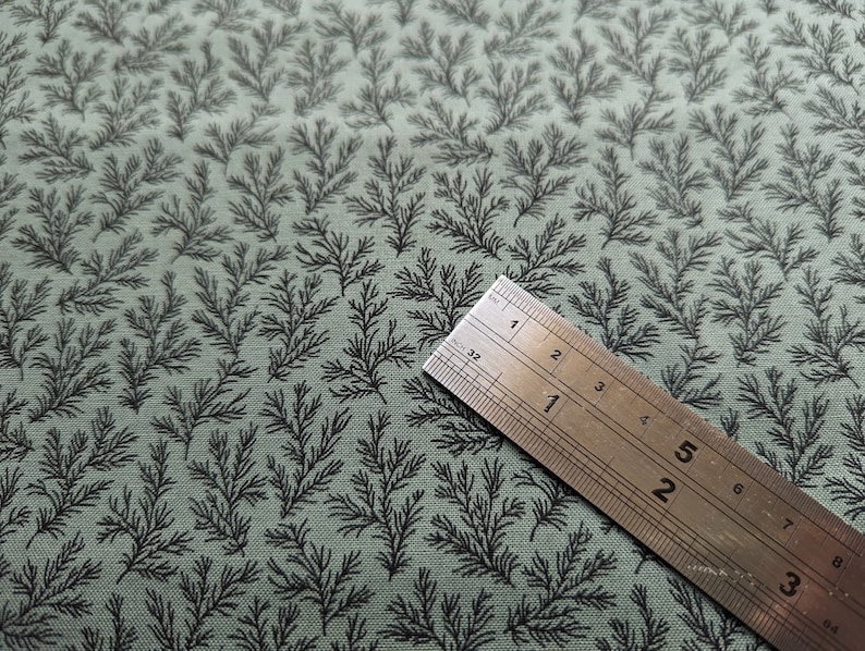 Pode incluir: Um close-up de um tecido verde com um padr&atilde;o repetitivo de pequenos ramos pretos estilizados. Uma r&eacute;gua de metal &eacute; colocada sobre o tecido, com os n&uacute;meros 1, 2, 3 e 4 vis&iacute;veis.