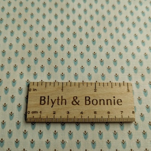 Puede incluir: Primer plano de tela con un patr&oacute;n repetido de formas florales turquesas y marrones sobre un fondo crema. Una regla de madera con el texto "Blyth & Bonnie" est&aacute; colocada sobre la tela. La regla mide hasta 20 cm.