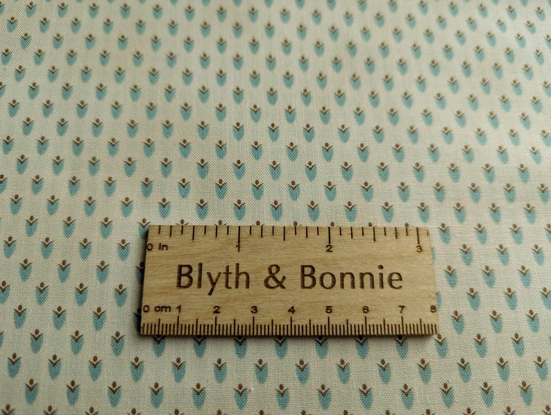 Puede incluir: Primer plano de tela con un patr&oacute;n repetido de formas florales turquesas y marrones sobre un fondo crema. Una regla de madera con el texto "Blyth & Bonnie" est&aacute; colocada sobre la tela. La regla mide hasta 20 cm.