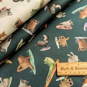 May include: A teal fabric with a repeating pattern of various animals, including a fox, owl, bear, and rabbit, all surfing on waves. The fabric features a ruler with the text "Blyth & Bonnie" printed on it.