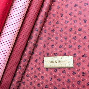 May include: Close-up of several fabric swatches in shades of red and pink. The fabrics feature various patterns, including small dots and floral designs. A wooden ruler with the text "Blyth & Bonnie" is at the bottom.