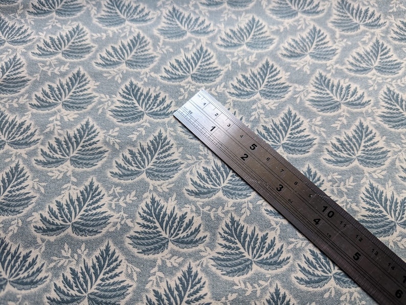 Pode incluir: Um close-up de um tecido azul e branco com um padr&atilde;o repetido de folhas de samambaia. Uma r&eacute;gua de metal &eacute; colocada em cima do tecido, mostrando medidas em polegadas.