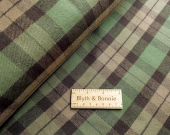 May include: Close-up of green, brown, and black plaid fabric. The fabric is folded over, revealing the texture and pattern. A wooden ruler with the text "Blyth & Bonnie" is placed on the fabric, showing measurements in inches.