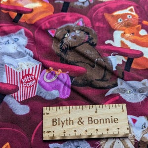 May include: A fabric featuring a repeating pattern of cats sitting in red theater seats, with one gray cat holding a red and white striped popcorn bucket labeled Kitty Corn. A wooden ruler measuring 3 inches is placed across the bottom of the fabric.