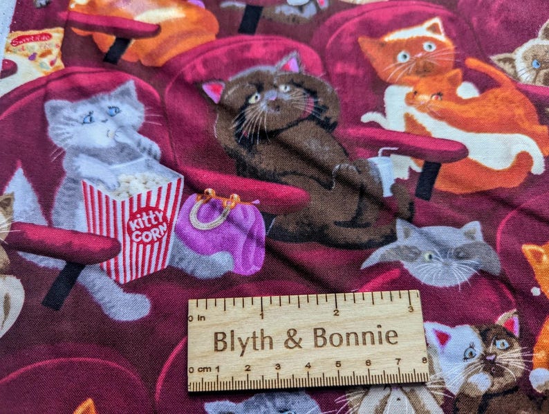 May include: A fabric featuring a repeating pattern of cats sitting in red theater seats, with one gray cat holding a red and white striped popcorn bucket labeled Kitty Corn. A wooden ruler measuring 3 inches is placed across the bottom of the fabric.
