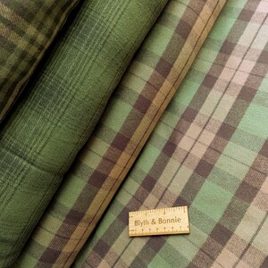 May include: Close-up of several rolls of plaid fabric in shades of green, brown, and tan. The fabric has a classic plaid pattern with intersecting lines. A wooden ruler with the text "Blyth & Bonnie" is visible.